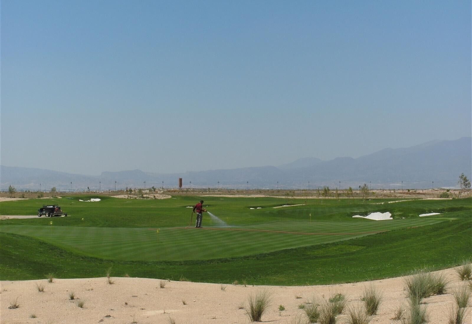 Un campo de golf con una persona regando el césped, un vehículo y dunas de arena al fondo.