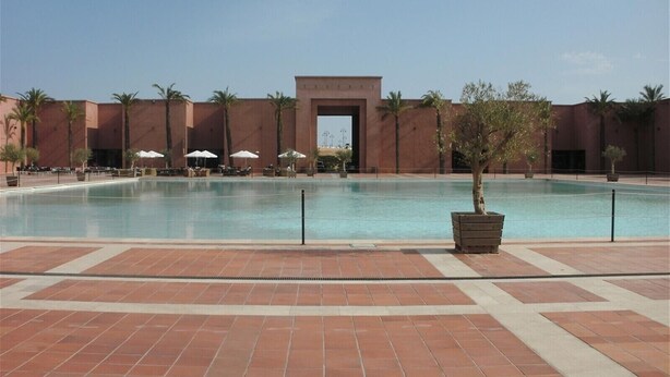 A large outdoor swimming pool with a tiled deck, palm trees, and a building with a central archway.