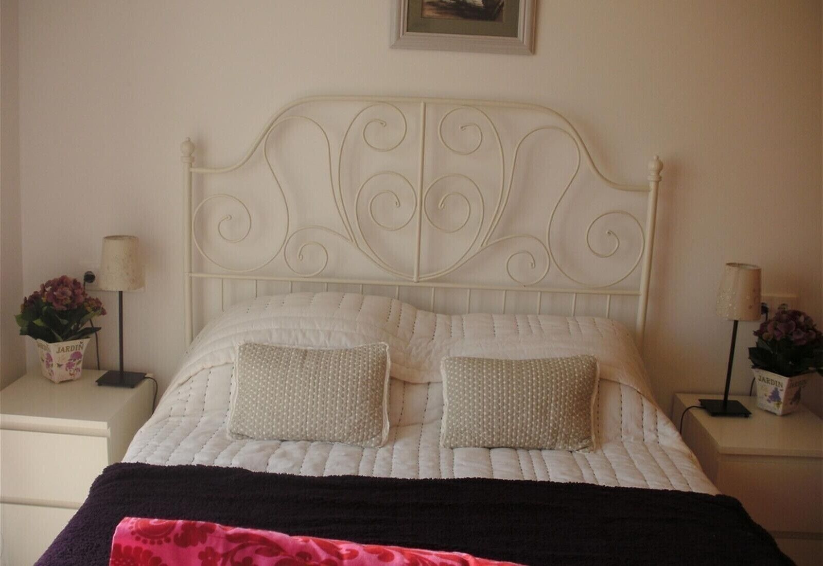 Una cama con cabecera blanca, dos almohadas, una colcha oscura y dos mesitas de noche con lámparas y jarrones de flores.
