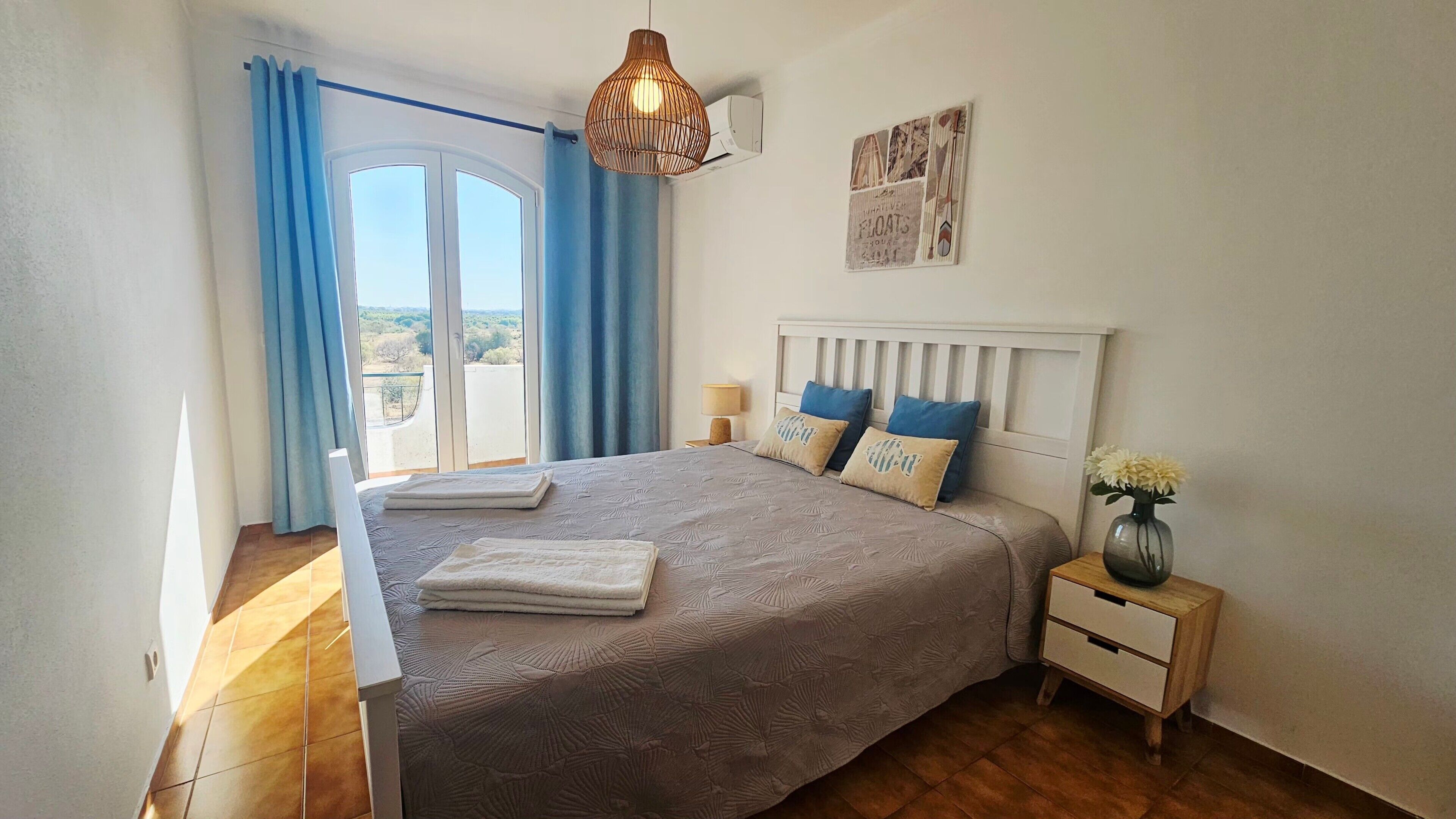 Een slaapkamer met een groot bed, een nachtkastje, een lamp, een vaas met bloemen en uitzicht op het landschap door een raam.