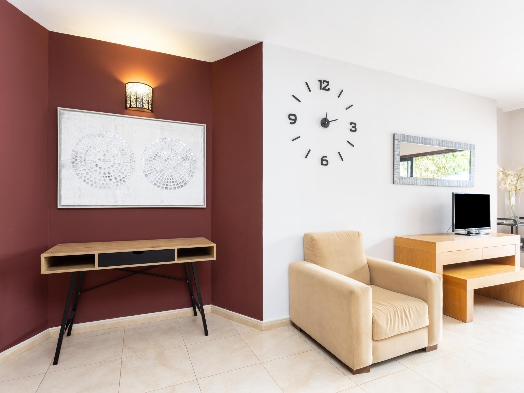 Un salon avec un mur rouge, une horloge blanche, une œuvre d’art encadrée, un fauteuil beige et un meuble TV en bois.