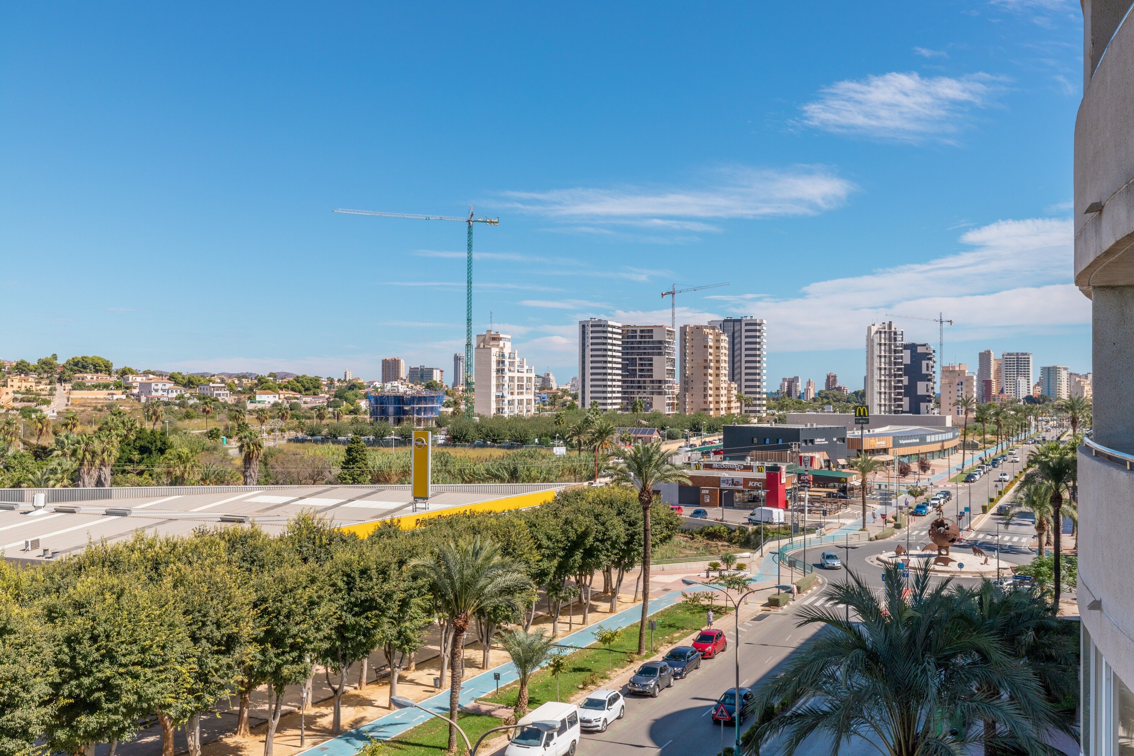 Un paisaje urbano con rascacielos, una grúa de construcción, palmeras y una calle concurrida con coches.