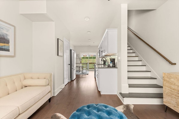 A modern interior with a sofa, a blue ottoman, a staircase, and a kitchen area.