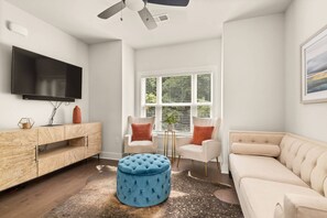 A modern living room with a flat-screen TV, a wooden cabinet, two white armchairs, a blue ottoman, and a beige sofa.