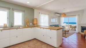 Una cucina moderna con mobili bianchi, un piano di lavoro in legno e vista sull'oceano attraverso ampie finestre.