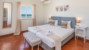 Un letto matrimoniale con testiera grigia, due comodini, una panca, un'ampia finestra vista mare e una lampada a soffitto.
