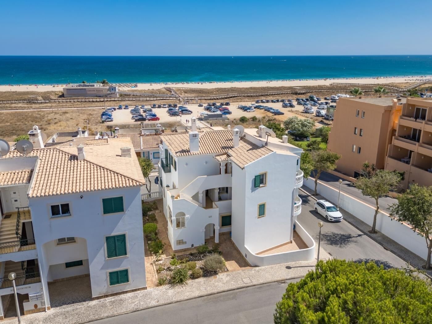 A coastal residential area with white buildings, a parking lot, and a beach in the background.