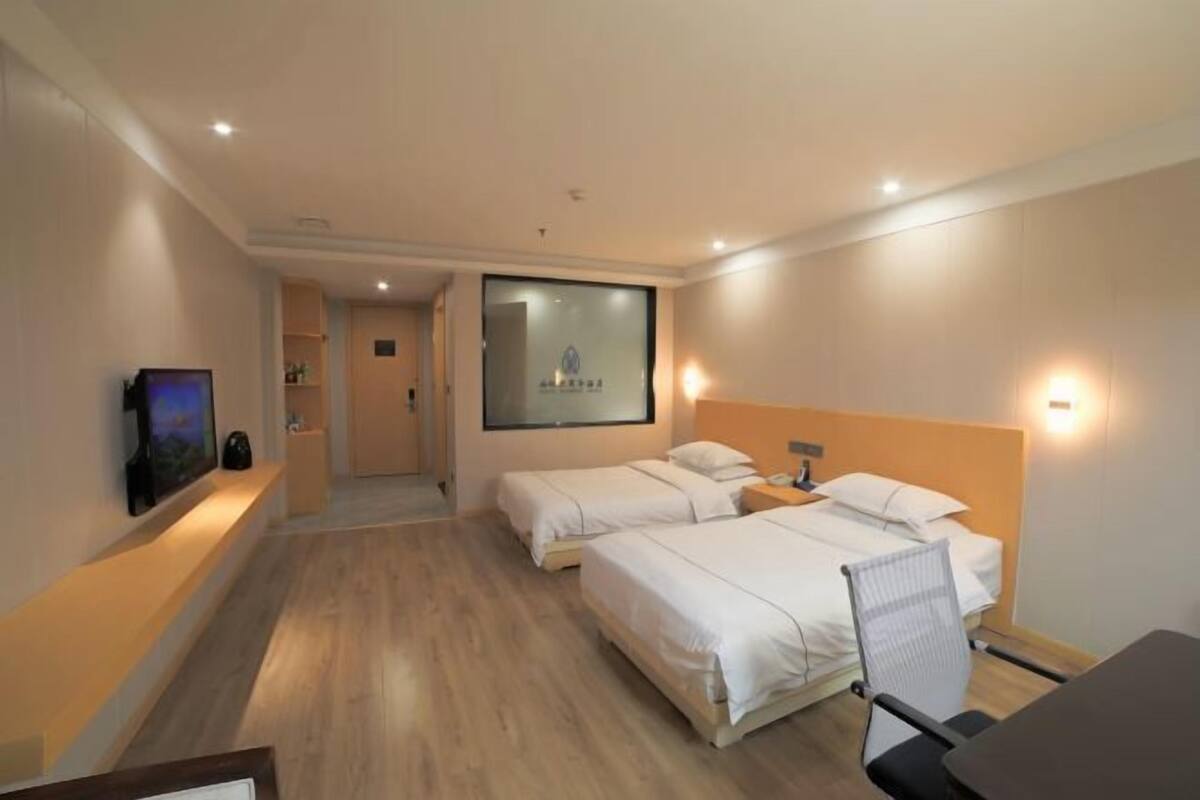 Habitación de hotel con dos camas, un escritorio, una silla, televisión y baño visible a través de una puerta de cristal.