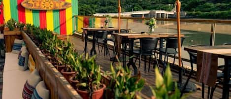 An outdoor terrace with tables and chairs, potted plants, and a view of a body of water.