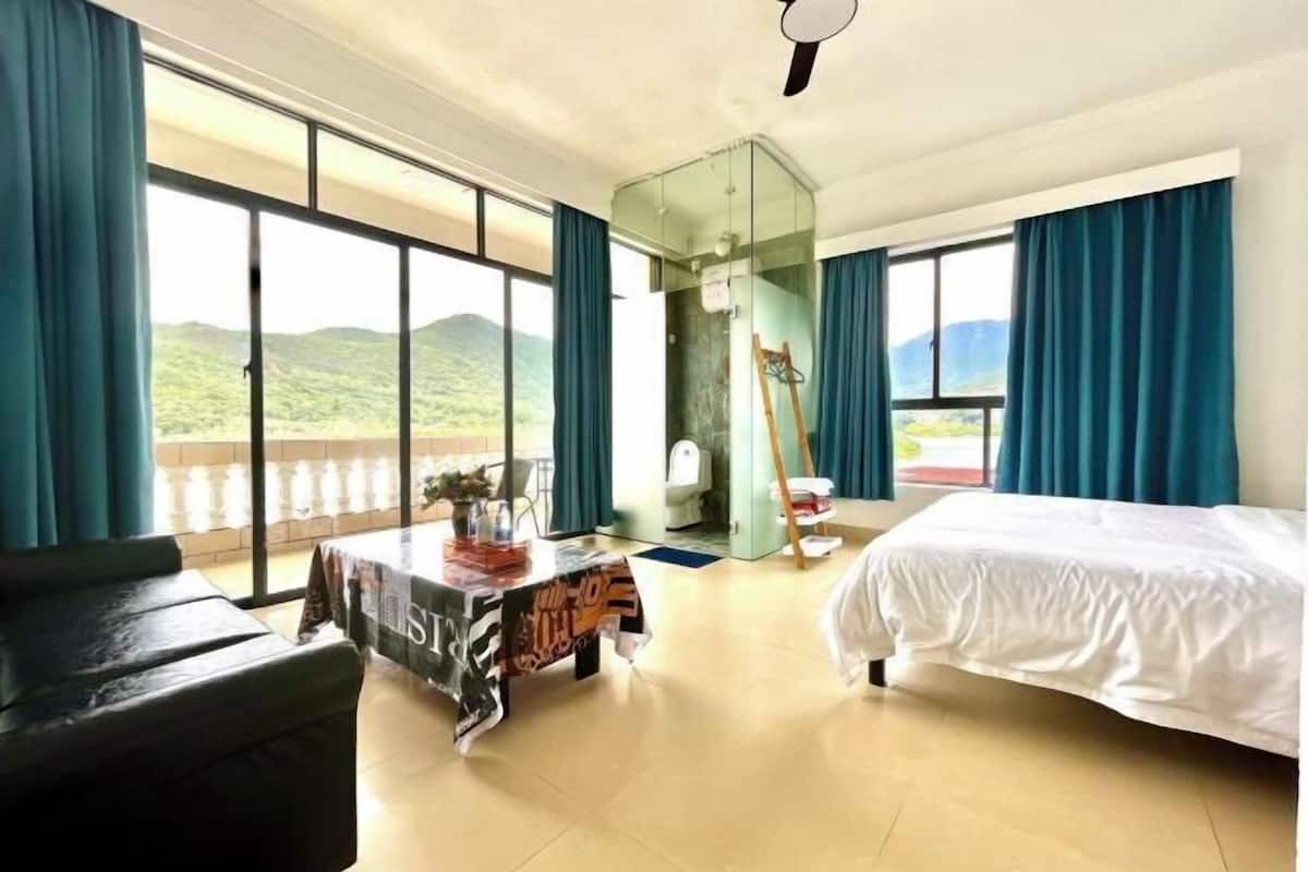 A hotel room with a bed, a table with a magazine, a chair, a glass partition, and a view of the outdoors.