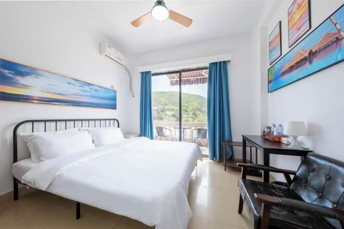 A hotel room with a bed, a desk with a chair, and a view of a mountainous landscape through a large window.