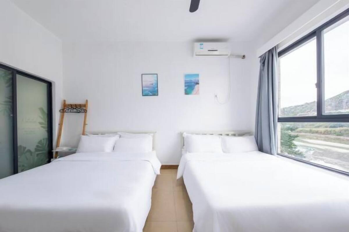 A hotel room with two beds, a window, a wall-mounted air conditioner, and a small shelf with a hanger.