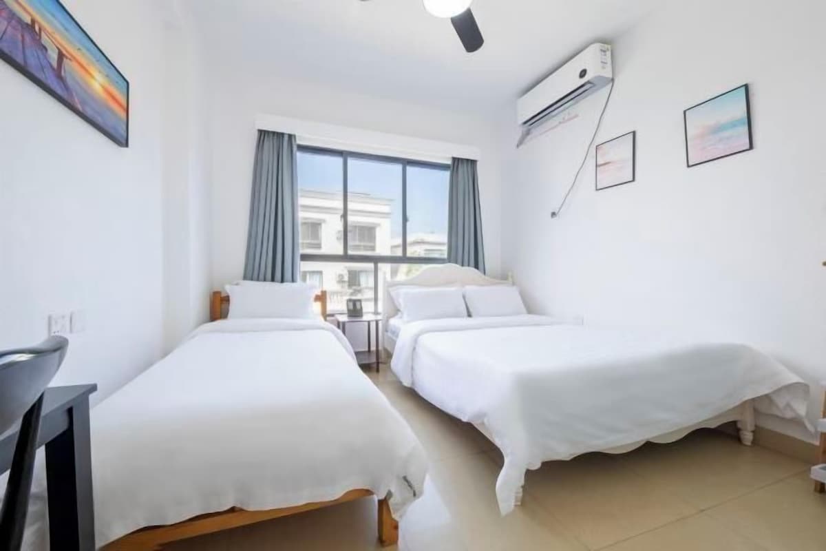 A hotel room with two single beds, a desk, a chair, a window with curtains, an air conditioning unit, and two framed pictures on the wall.