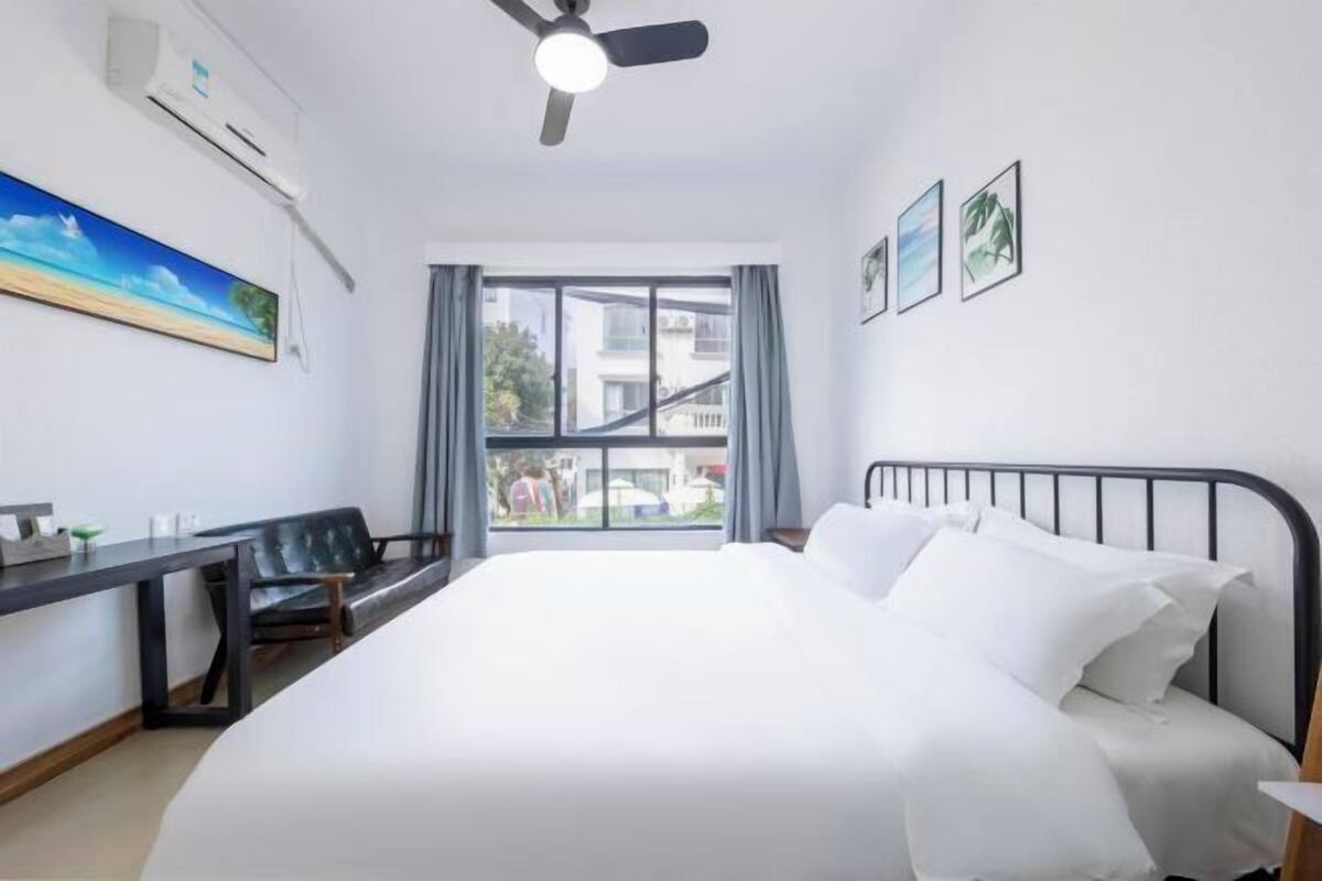 A hotel room with a bed, desk, chair, ceiling fan, and large window.