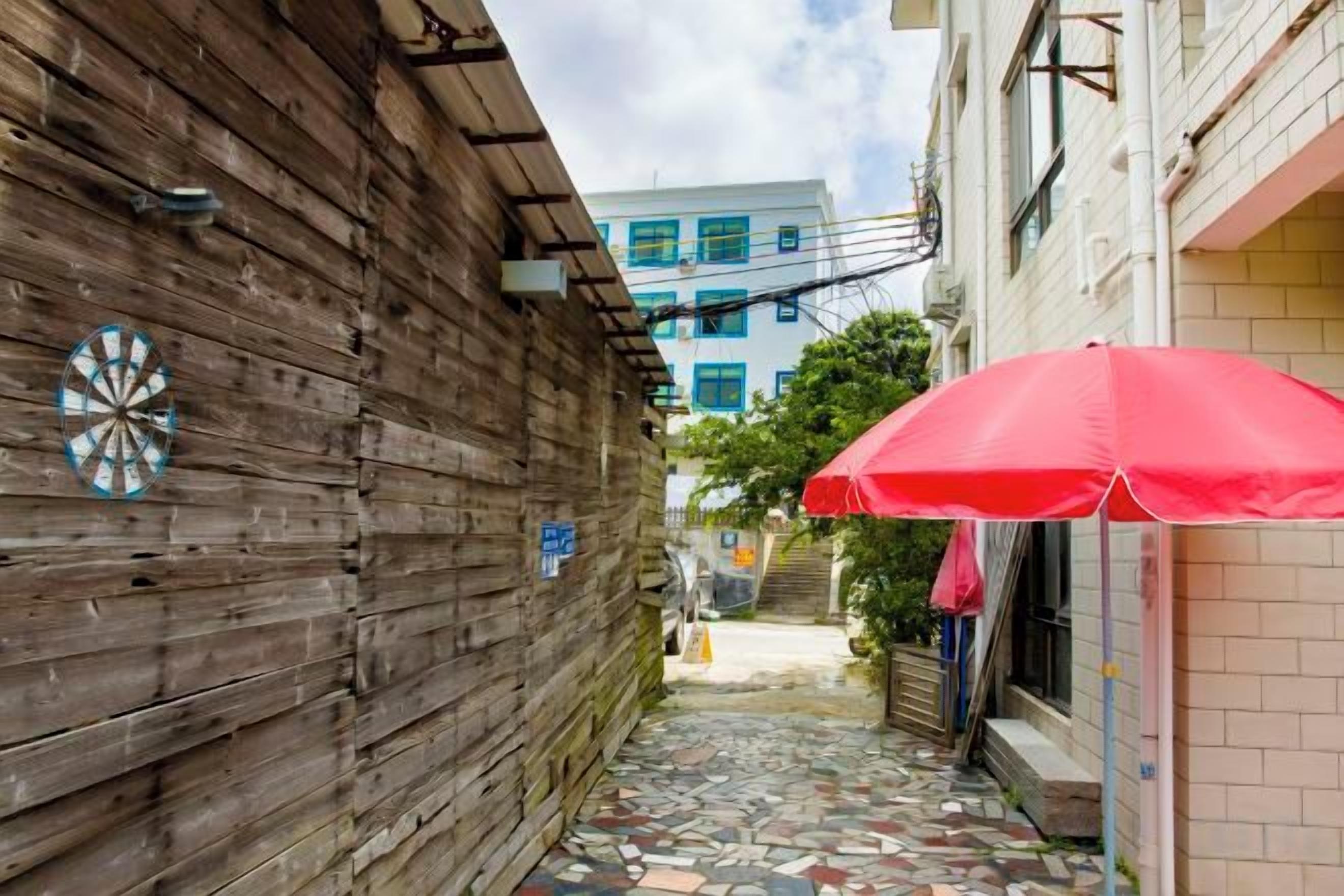 A narrow alleyway with a wooden wall, a red umbrella, and a building with a tiled floor in the background.