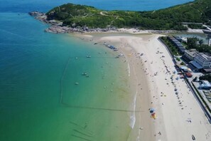 An aerial view of a beach with clear waters, a sandy shore, and a small hill with vegetation.