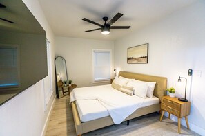 A bedroom with a bed, bedside table, nightstand, ceiling fan, and a window with blinds.