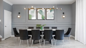 Un espace repas avec une table rectangulaire entourée de six chaises noires, un lustre doté de plusieurs points lumineux et des œuvres d’art encadrées accrochées au mur.
