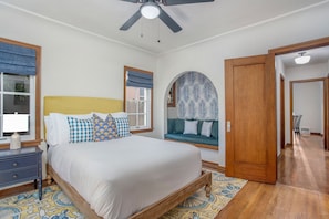 A bedroom with a bed, a nightstand, a window with blue curtains, a ceiling fan, and a wooden door. - Villa La Verne (Long Beach)
