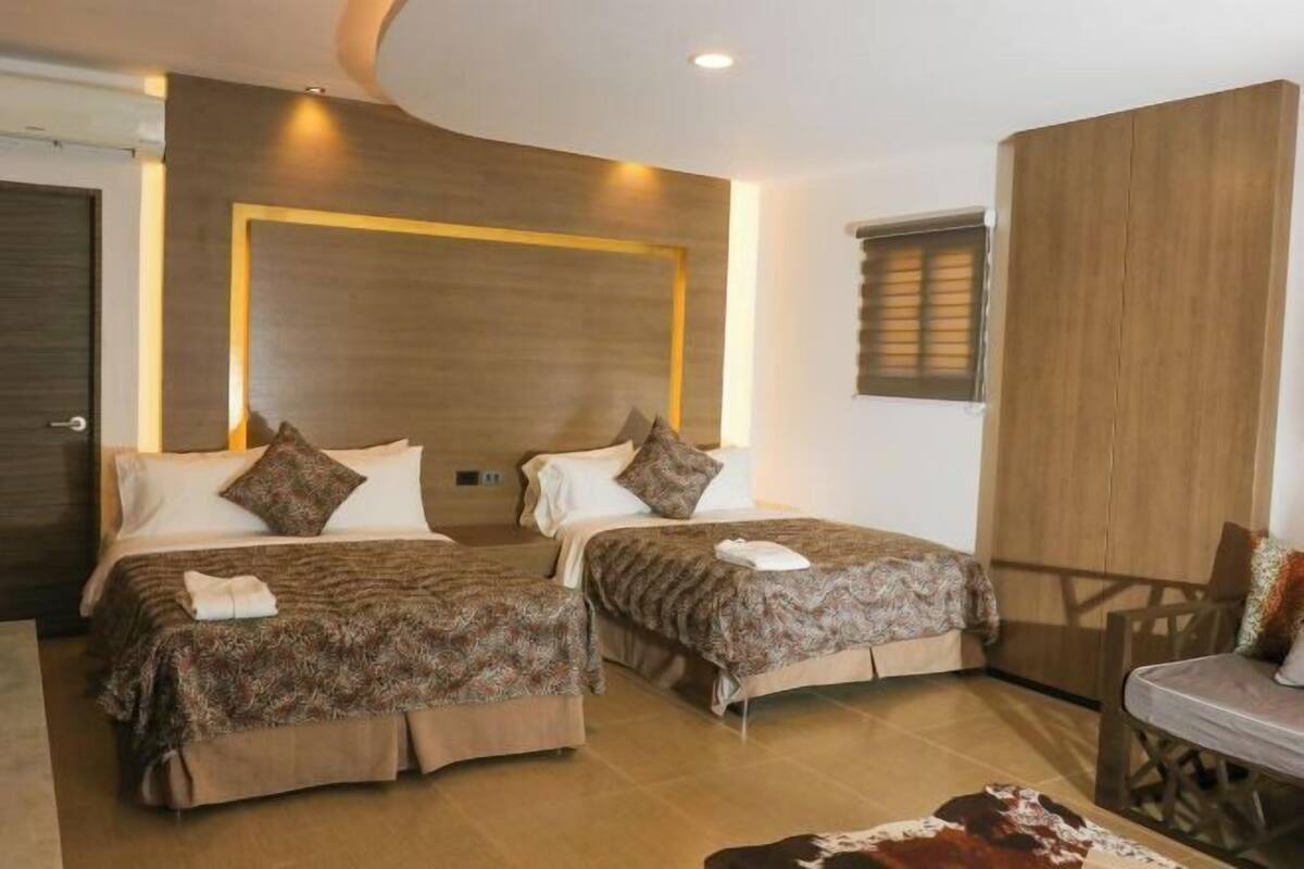 Habitación de hotel con dos camas, cabecera de madera, una mesita pequeña y un tapete de cuero de vaca.
