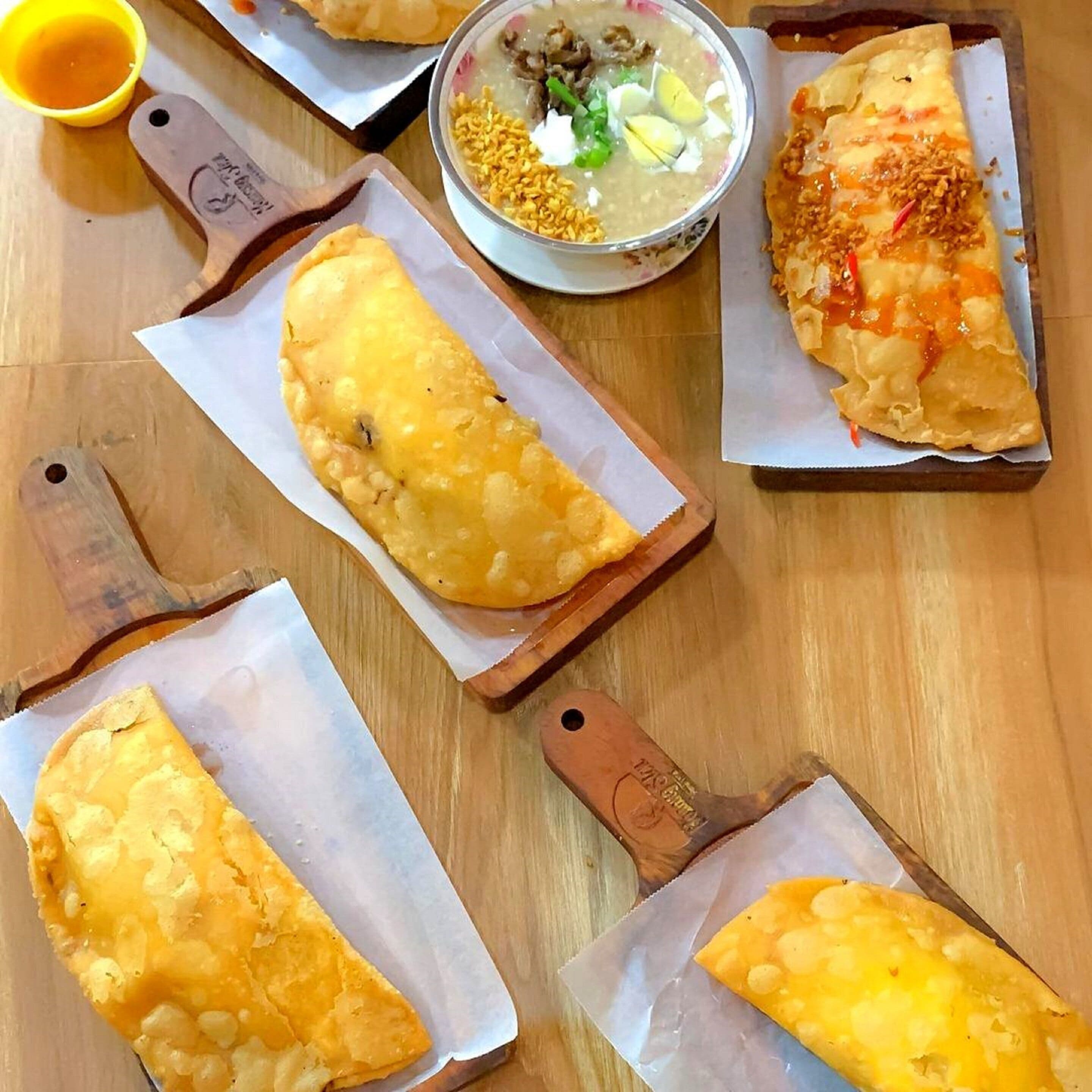 A wooden table with three food items: two triangular fried pastries on wooden boards and a bowl of soup with noodles and vegetables.