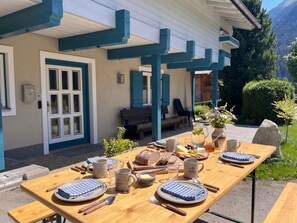 An outdoor dining area with a wooden table set for a meal, a bench, and a house with blue accents. - Group house overlooking the waterfalls (Krimml)