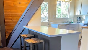 Cucina con soffitto in legno, un piano di lavoro bianco, due sgabelli da bar e una finestra vista verde.