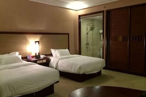 Habitación de hotel con dos camas, una mesita de noche con lámpara, zona de ducha y una mesa redonda.