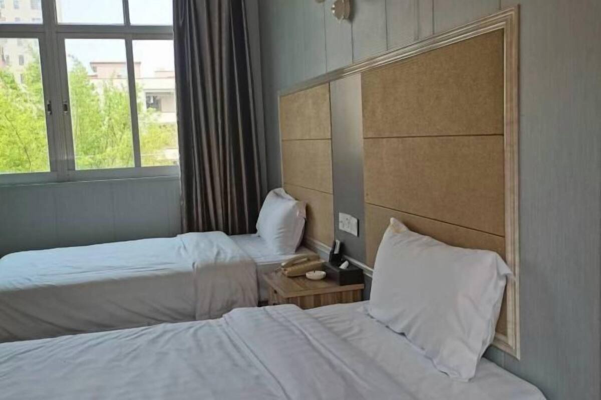 Een hotelkamer met twee bedden, een houten hoofdbord, een nachtkastje met een lamp en een raam met gordijnen.