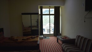 A room with a bed, a sofa, a small table, a window with curtains, and a view of a mountainous area. - Hotel Cedar Inn (Mansehra)