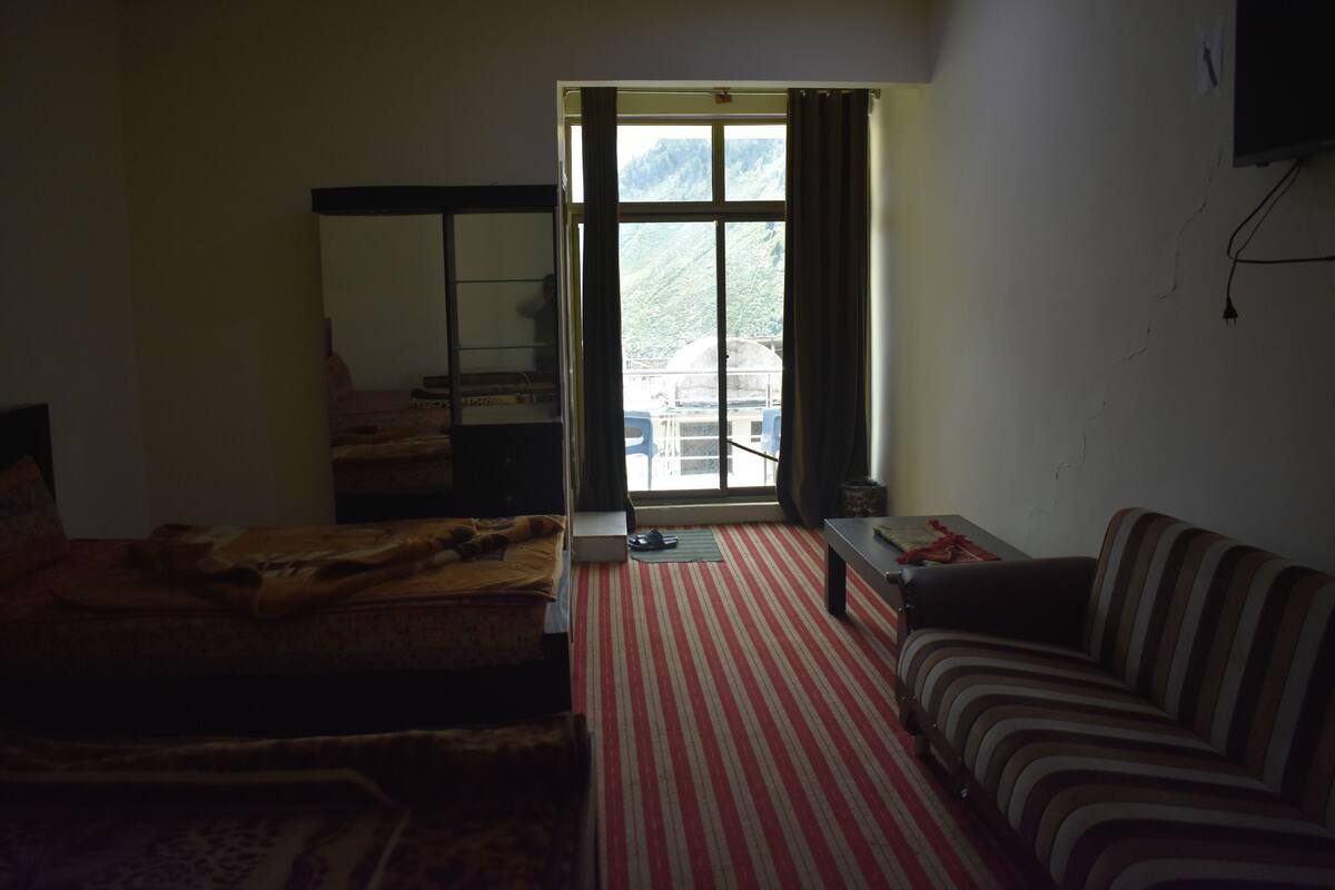 A room with a bed, a sofa, a small table, a window with curtains, and a view of a mountainous area.