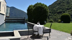 Zona pranzo a bordo piscina con un tavolo apparecchiato per due, immersa in un prato curato e con le montagne sullo sfondo.