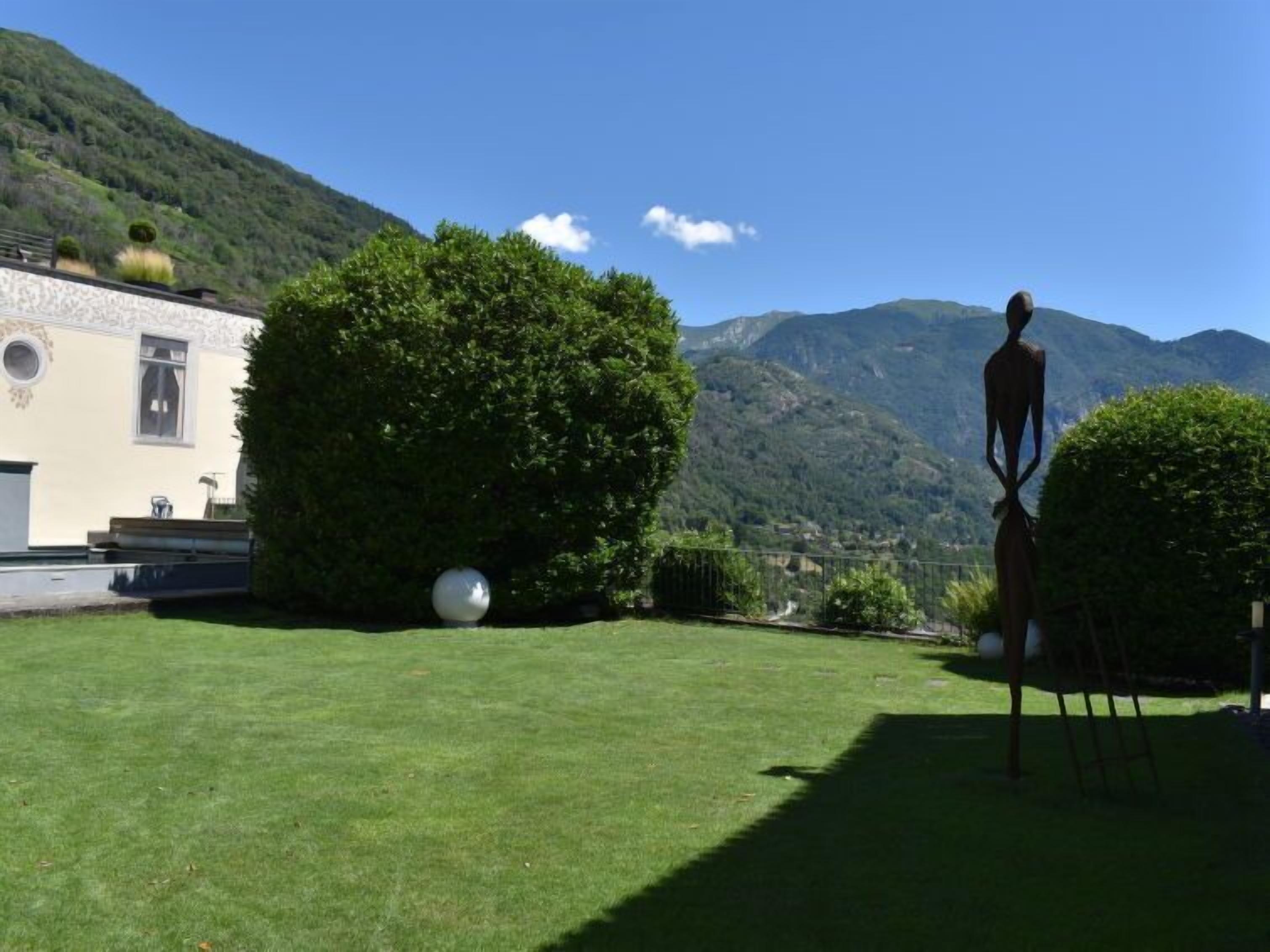 A grassy courtyard with a sculpture, a building with a white facade, and mountains in the background.