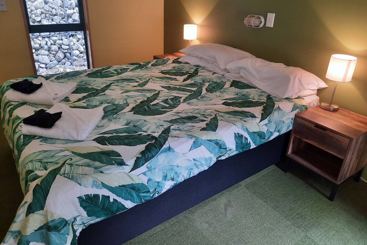 A bed with a tropical leaf-patterned bedspread, two towels, two bedside lamps, and a nightstand with a lamp.