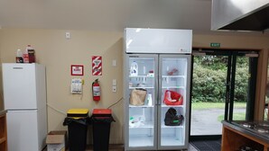 A kitchen area with a refrigerator, a freezer, and a double-door icebox. There are two trash bins, a fire blanket, and a fire extinguisher on the wall.