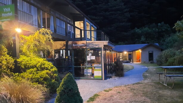 A reception area with a glass door, a sign reading "YHA Hostel," and a small building in the background.