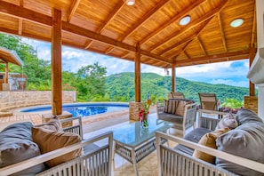 Un espacio al aire libre techado con piscina, mobiliario y vistas a un paisaje montañoso.