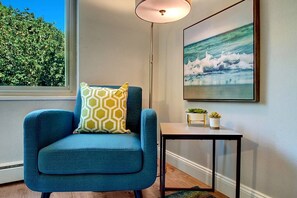 A blue armchair with a geometric patterned pillow, a small table with potted plants, and a framed picture of ocean waves on the wall.