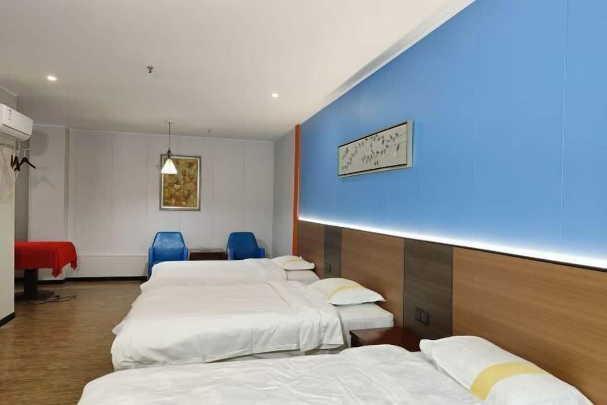 Quarto de hotel com três camas, uma mesa vermelha, uma cadeira azul e uma obra de arte pendurada na parede.