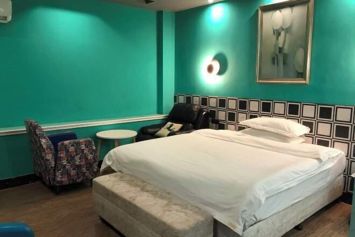 Quarto de hotel com paredes verde-água, cama com roupa de cama branca, cabeceira preta, poltrona estampada e uma pequena mesa redonda.