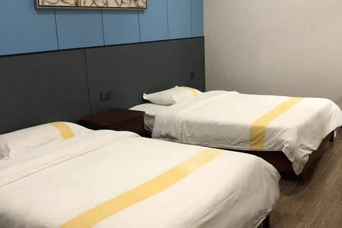 Quarto de hotel com duas camas, cada uma com roupa de cama branca e uma faixa amarela. O quarto tem parede azul e piso de madeira.