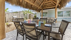 An outdoor patio with a thatched roof, wooden chairs, a table set with plates and glasses, and a view of a house in the background. - Heated Pool, Tiki Hut, Game Room, Beach Gear, BBQ! (Cocoa Beach)