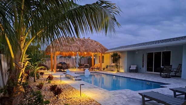 A pool area with lounge chairs, a thatched-roof structure, and a stone pathway. - Heated Pool, Tiki Hut, Game Room, Beach Gear, BBQ! (Cocoa Beach)