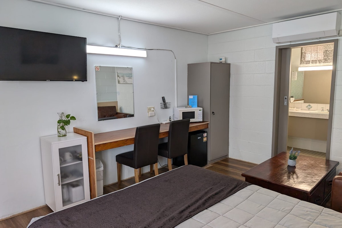 A compact hotel room with a bed, a desk with two chairs, a microwave, a small cabinet, and a television mounted on the wall.