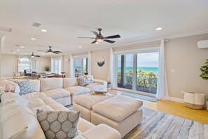 Ein geräumiges Wohnzimmer mit einer großen Eckcouch, mehreren Deckenventilatoren und Blick auf das Meer durch große Schiebetüren.