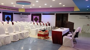 Un salón de conferencias con filas de sillas cubiertas de blanco, un escenario con una mesa roja y un equipo de aire acondicionado en la pared.