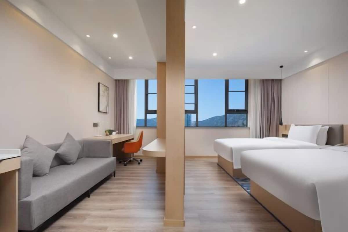 A hotel room with a sofa, a desk, two beds, and a large window with a view.
