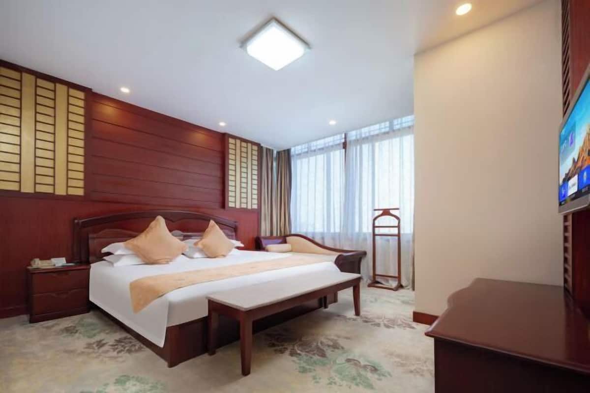 A hotel room with a wooden headboard, a large bed with multiple pillows, a bedside table, a bench, and a TV on a stand.