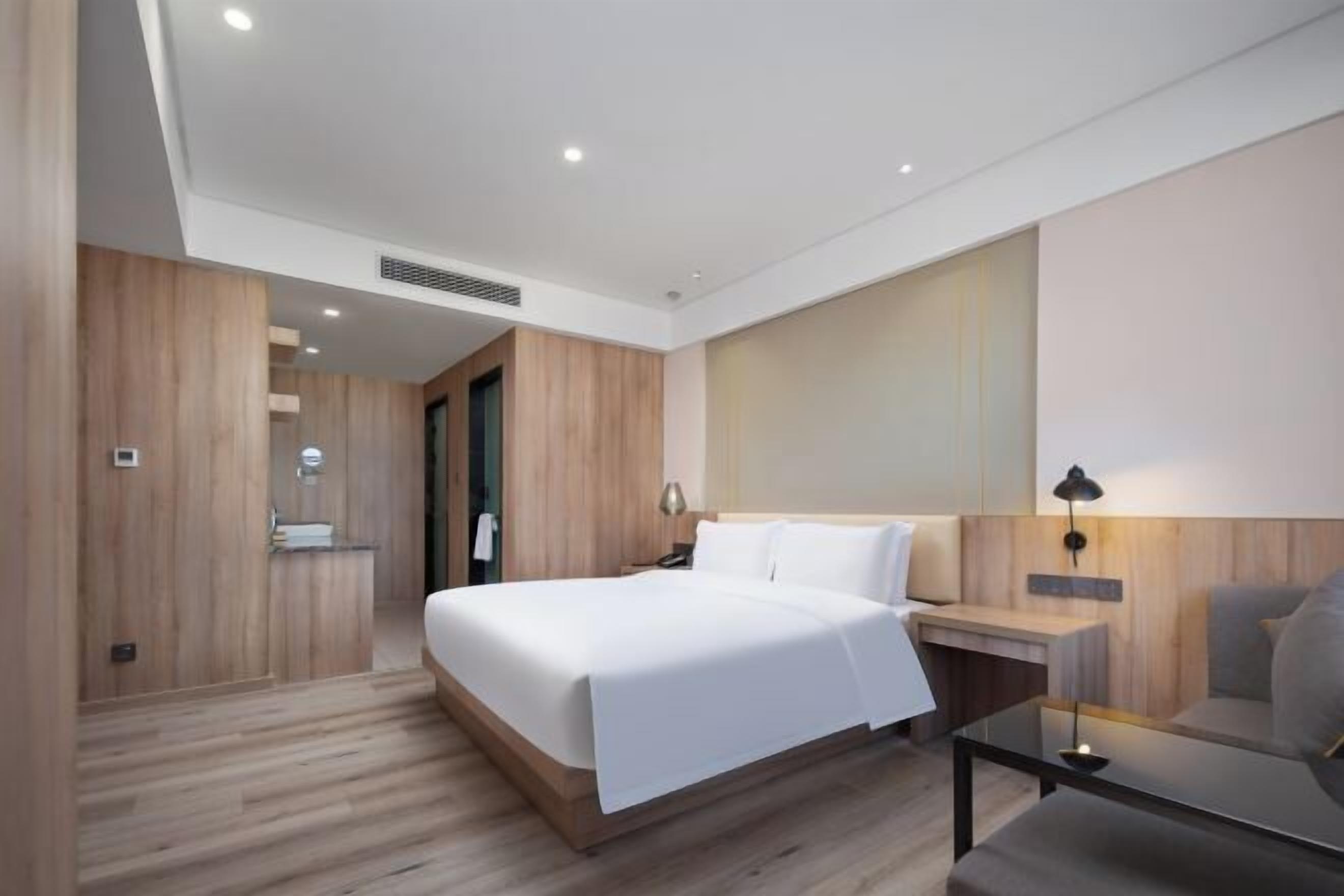 A modern hotel room with a large bed, a bedside table, a desk, and a chair.