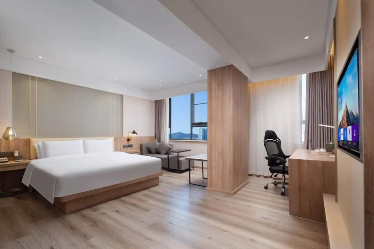 A modern hotel room with a large bed, a desk, a chair, a TV, and a window with a city view.